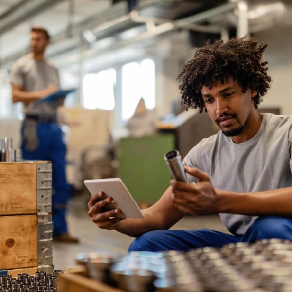 African American manual worker using touchpad while examining manufactured stainless steel rods in a factory.